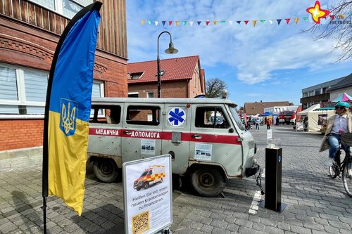 Ein beschossener, grauer, alter Rettungswagen steht auf einer Straße in Langenberg. Im Vordergrung ist ein Aufsteller mit ukrainischer Flagge zu sehen. Eine Radfahrerin fährt vorbei. In der Luft hängt eine Wimpelkette.