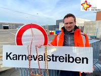 Bauhofleiter Jens Hökenschnieder steht auf dem Außengelände des Bauhofs und zeigt ein Straßenschild auf dem das Wort Karnevalstreiben zu lesen ist. Im Hintergund ist ein weiteres Straßenschild zu sehen. Das Foto ist oben rechts mit dem Logo der Stadt Rietberg gekennzeichnet.