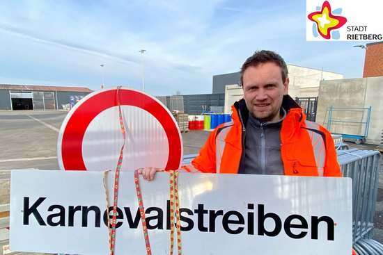 Bauhofleiter Jens Hökenschnieder steht auf dem Außengelände des Bauhofs und zeigt ein Straßenschild auf dem das Wort Karnevalstreiben zu lesen ist. Im Hintergund ist ein weiteres Straßenschild zu sehen. Das Foto ist oben rechts mit dem Logo der Stadt Rietberg gekennzeichnet.