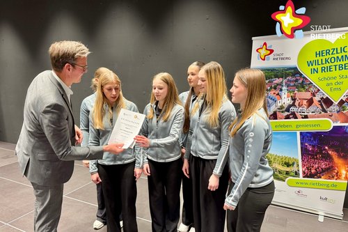 Bürgermeister Andreas Sunder im grauen Anzug überreicht auf einer Bühne eine Urkunde an sortliche gekleidete junge Frauen.