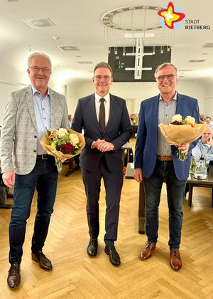 Drei Männer stehen lächelnd nebeneinander, die zwei außenstehenden halten Blumen in den Händen.