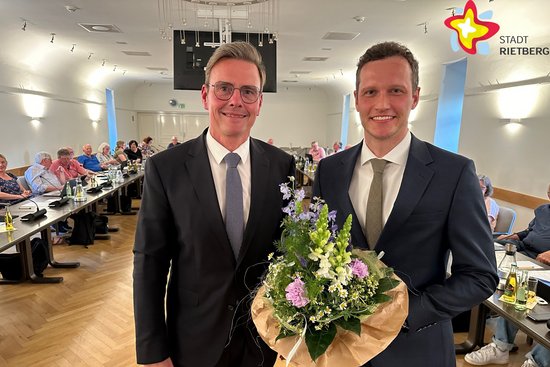 Bürgermeister Andreas Sunder und Florian Kapp stehen im Ratssaal uns schauen in die Kamera. Herr Kapp hält einen Blumenstrauß in der Hand. Im Hintergrund sind sitzende Ratsmitglieder zu sehen.