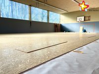 Blick in die Turnhalle. Ein Holzboden ist zum Teil bereits verlegt, ein kleiner Teil ist noch mit Folie bedeckt.