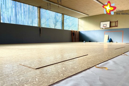 Blick in die Turnhalle. Ein Holzboden ist zum Teil bereits verlegt, ein kleiner Teil ist noch mit Folie bedeckt.