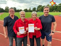 Brigitte und Ferdi Schumacher stehen auf der Laufbahn des Sportplatzes und halten Urkunden in den Händen. Eingerahmt werden sie von Meinolf Hillemeier und Bürgermeister Andreas Sunder.