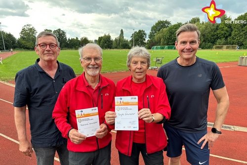 Brigitte und Ferdi Schumacher stehen auf der Laufbahn des Sportplatzes und halten Urkunden in den Händen. Eingerahmt werden sie von  Meinolf Hillemeier und Bürgermeister Andreas Sunder.