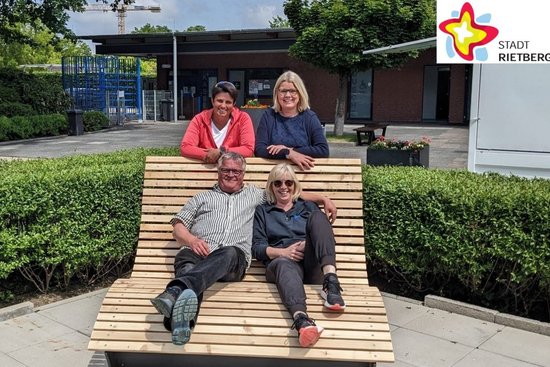 Andreas Pauleickhoff und Freibad-Chefin Kirsten Meier sitzen im Freibad auf einer überdimensionalen Liege, die mit dhellen dünnen Holzlatten versehen ist. Schwimmmeisterin Birgit Neugebauer und Doris Schulz von der städtischen Abteilung Schule, Kultur und Sport stehen lächelnd dahinter.