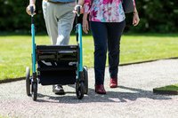 Hinter einem Rollator auf einem Fußwege neben einem grüen Rasen sind die Beine zweier Menschen zu sehen.