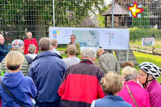 Vor mehreren an einem Fußballtor befestigen Bauplänen spricht Bürgermeister Andreas Sunder vor mehreren Menschen, die lediglich von hinten zu sehen sind.