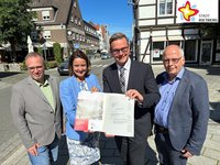 Drei Männer und eine Frau stehen draußen auf der Straße und halten einen Förderbescheid in die Kamera. Im Hintergrund sind Häuser und blauer Himmel zu sehen.