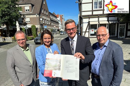 Drei Männer und eine Frau stehen draußen auf der Straße und halten einen Förderbescheid in die Kamera. Im Hintergrund sind Häuser und blauer Himmel zu sehen.