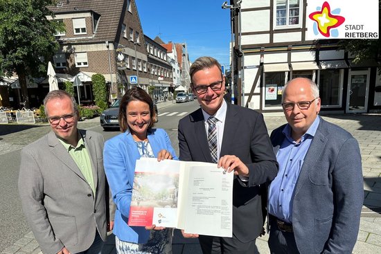 Drei Männer und eine Frau stehen draußen auf der Straße und halten einen Förderbescheid in die Kamera. Im Hintergrund sind Häuser und blauer Himmel zu sehen.