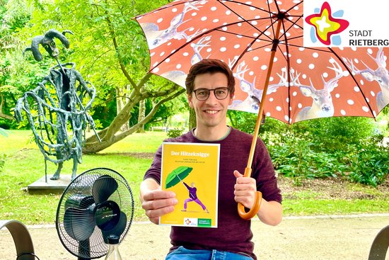 Lukas Schmidt, Klimaanpassungsmanager der Stadt Rietberg, sitzt im Klostergarten auf einer Bank und hält einen Schirm über sich sowie eine Broschüre in der linken Hand. Rechts neben ihm stehen ein Ventilator und eine Flasche Wasser.