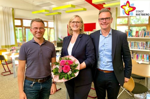 Dominik Bartels und Andreas Sunder stehen mit Birgit Schneider, die einen Blumenstrauß hält, in der Schulbibliothek des Grundschulstandortes Westerwiehe.