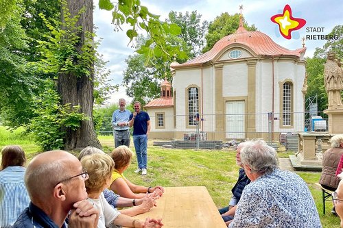 Vor der strahlend weißen Johanneskapelle stehen Michael Kaune und Andreas Sunder. Im Vordergrund sitzen Menschen an einem leeren Tisch.