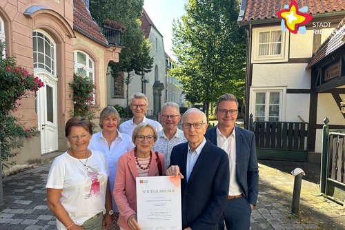 Die genannten Personen stehen zwischen Fachwerkhäusern, im Hintergrund ist die Klosterkirche zu sehen. Sie schauen in die Kamera.
