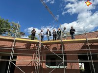 Die genannten Personen stehen auf einem Gerüst des Neubaus und schauen nach unten. Im Hintergrund weht ein Richtkranz mit bunten Bändern.
