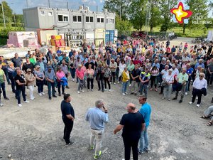 Hunderte Frauen und Männer stehen auf einer Baustelle im Halbkreis um weitere vier Männer, die in etwas Abstand und mit dem Rücken zur Kamera stehen.