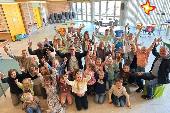 Die Kinder und die genannten Personen sitzen in der neuen Aula auf dem Fußboden und strecken begeistert die Hände in die Luft. Sie schauen in die Kamera.