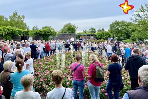 Dutzende Frauen und Männer stehen mit dem Rücken zur Kamera im Kreis um ein Blumenbeet und lauschen einem Redner im Hintergrund.