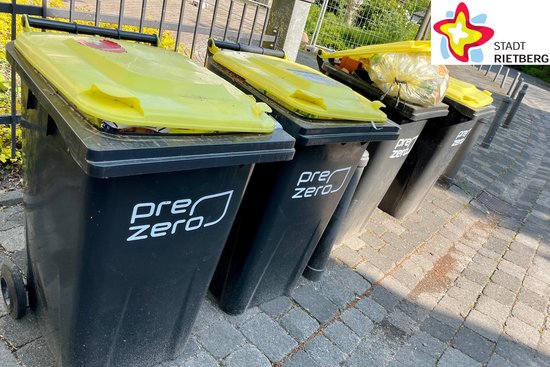 Vie graue Tonnen mit gelben Deckeln stehen nebeneinander am Straßenrand. Bei einer der Tonnen lässt sich der Deckel nicht schließen, weil sich darin der Müll so hoch aufgetürmt hat.