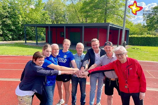 Monika Nordmann (Finanzvorstand bei TuS Viktoria Rietberg), Franziska Rehkemper (Abteilungsleiterin Leichtathletik beim TuS), Ferdinand Schumacher (stellvertretender Vorsitzender des Stadtsportverbandes), Lisa Schlüter (stellv. Abteilungsleiterin Leichtathletik), Stadtsportverband-Jugendwart Hansjosef Stücker, Bürgermeister Andreas Sunder, Zimmermann Sebastian Hiebing und Sportabzeichenobmann Willi Wilma stehen im Halbkreis und reichen sich je eine Hand in der Bildmitte.