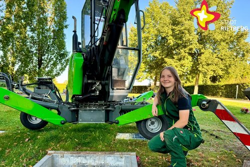 Eine junge Frau in grüner Arbeitskleidung kniet auf einer grünen Wiese hinter einem gerade ausgehobenen Grab und vor einem speziellen Friedhofsbagger