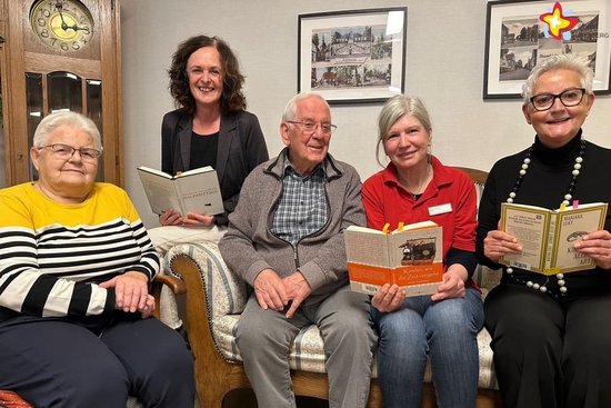 Mitarbeiterin und Lesepatin aus der Stadtbibliothek sitzen gemeinsam mit Caritas-Mitarbeiterin und Gästen der Tagespflege auf dem Sofa und halten die Bücher in Händen, aus denen vorgelesen wird.