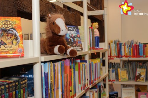 Zu sehen sind Regale mit Kinderliteratur, oben auf einem der Regale sitzt ein Stofftier-Pferd.