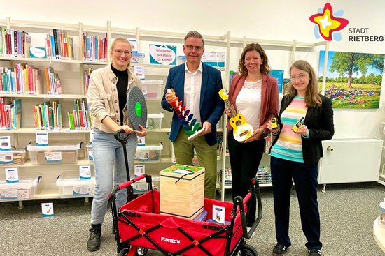 Wibke Kordtomeikel, Bürgermeister Andreas Sunder und Julia Bröckling und Jennifer Bader als Leitungsteam der Stadtbibliothek stehen hinter einem Bollerwagen und halten ein Balance Board, einen Klangbaum, eine Ukulele und zwei Walkie Talkies in den Händen.