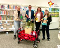 Wibke Kordtomeikel, Bürgermeister Andreas Sunder und Julia Bröckling und Jennifer Bader als Leitungsteam der Stadtbibliothek stehen hinter einem Bollerwagen und halten ein Balance Board, einen Klangbaum, eine Ukulele und zwei Walkie Talkies in den Händen.