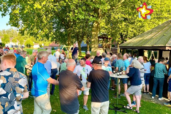 Auf grünem Gas und rechts von einer hölzernen Grillhütte stehen viele Menschen eng beeinander, teils auf einen Stehtisch gelehnt.