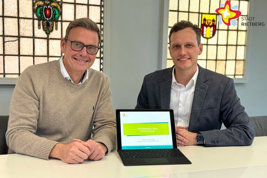 Die beiden Genannten sitzen in einem BEsprechungsraum und schauen in die Kamera. Zwischen ihnen steht ein aufgeklappter Laptop.