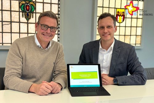 Die genannten Personen sitzen nebeneinander an einem Tisch, schauen in die Kamera. Zwischen ihnen steht ein kleiner Laptop.