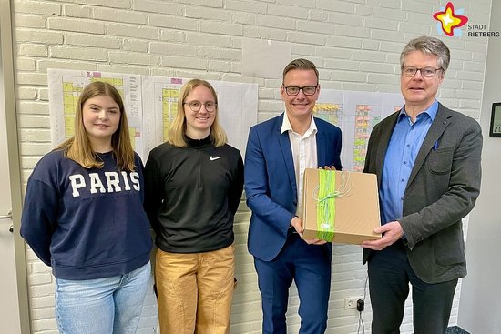 Zwei Schülerinnen stehen neben dem Bürgermeister, der gemeinsam mit dem neuen Schulleiter einen Geschenkkarton in den Händen hält.