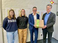 Zwei Schülerinnen stehen neben dem Bürgermeister, der gemeinsam mit dem neuen Schulleiter einen Geschenkkarton in den Händen hält.