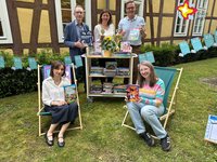 Die genannten Personen sind im Garten. Zwei sitzen auf Liegestühlen, drei stehen hinter einem kleinen Bücherregal. Sie schauen in die Kamera.