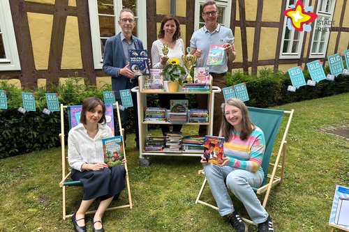 Die genannten Personen sind im Garten. Zwei sitzen auf Liegestühlen, drei stehen hinter einem kleinen Bücherregal. Sie schauen in die Kamera.