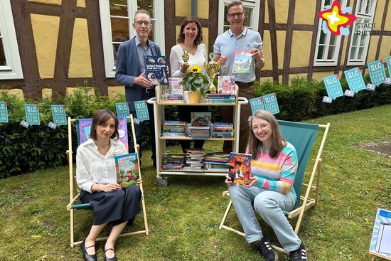 Die genannten Personen sind im Garten. Zwei sitzen auf Liegestühlen, drei stehen hinter einem kleinen Bücherregal. Sie schauen in die Kamera.