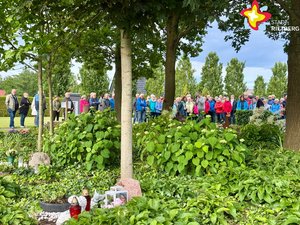 Mehrere Menschen stehen hinter einem grünen Beet, im Vordergund ist eine Grabstelle mit Kerze zu sehen