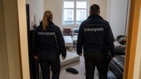 Zwei Mitarbeiter eines Ordnungsamts sind von hinten zu sehen. Sie stehen in einem nicht aufgeräumten Zimmer.
