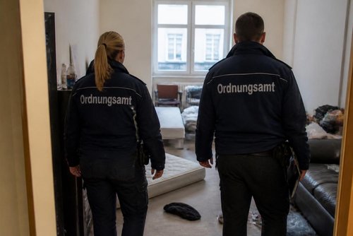 Zwei Mitarbeiter eines Ordnungsamts sind von hinten zu sehen. Sie stehen in einem nicht aufgeräumten Zimmer.
