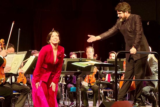 Sopranistin Anne Elizabeth Sorbara in einem roßten Hosenanzug und bekommt Applaus vom Dirigenten Markus Huber.