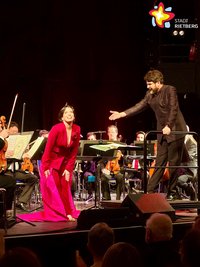 Sopranistin Anne Elizabeth Sorbara in einem roßten Hosenanzug und bekommt Applaus vom Dirigenten Markus Huber.