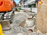 In einer Baustelle sind ein Bagger und ein Kanalschaft zwischen reichlich Schotter und Kies zu sehen.