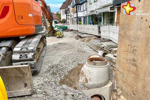 In einer Baustelle sind ein Bagger und ein Kanalschaft zwischen reichlich Schotter und Kies zu sehen.