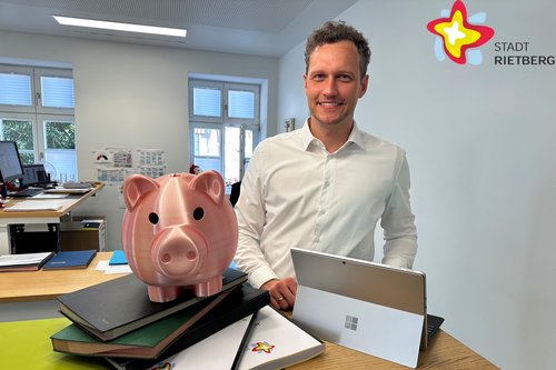Florian Kapp steht an einer Theke im Vorzimmer im Rathaus. Neben ihm steht ein rosafarbenes Sparschwein. Akten liegen auf der Theke.