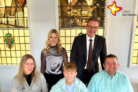 Ein Junge sitzt hinter einem großen, aufgeschlagenen Buch am Tisch, links und rechts sitzen Mutter und Vater. Dahinter stehen eine junge Frau sowie ein Bürgermeister im Anzug.