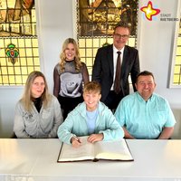 Ein Junge sitzt hinter einem großen, aufgeschlagenen Buch am Tisch, links und rechts sitzen Mutter und Vater. Dahinter stehen eine junge Frau sowie ein Bürgermeister im Anzug.