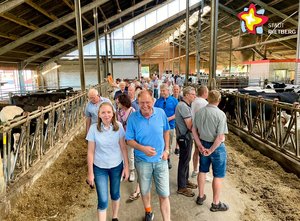 Mehrere Menschen laufen hintereinander durch einen Stall; links und rechts sind Kühe zu sehen.